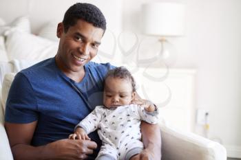 Close up of young adult black father sitting in an armchair holding his three month old baby son, smiling to camera, front view