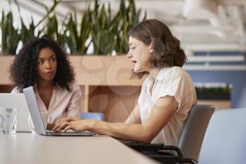 Two Businesswomen Working On Laptops Sitting At Table In Modern Open Plan Office