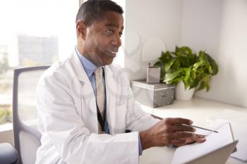 Male Doctor Wearing White Coat In Office Sitting At Desk Working On Laptop
