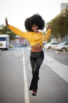 Young black woman with afro jumps, looking to camera, while crossing street, front view, vertical
