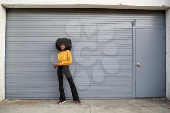 Hip young black woman standing against grey security shutters smiling to camera, full length