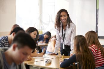 Portrait Of Smiling Female High School Teacher Standing By Student Table Teaching Lesson