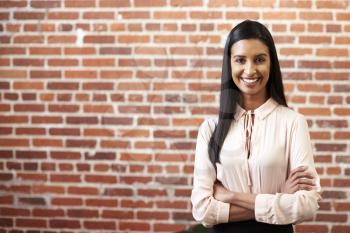 Portrait Of Smiling Young Businesswoman Standing Against Brick Wall In Modern Office