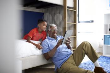 Grandfather With Grandson Lying In Bedroom Playing Game On Digital Tablet Together