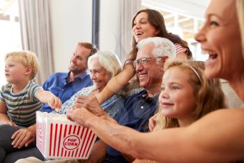 Multi-Generation Family Sitting On Sofa At Home Eating Popcorn And Watching Movie Together