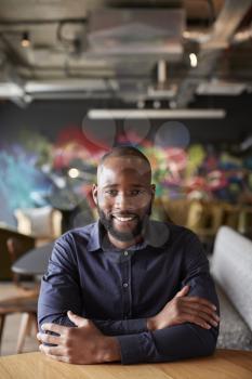 Mid adult black male creative sitting at a table with arms crossed smiling to camera, close up