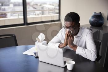 Young black businessman sitting at an office desk blowing his nose into a tissue, elevated view