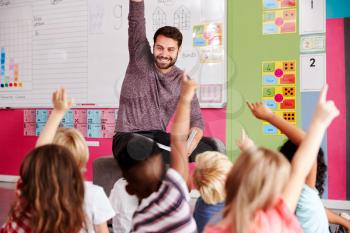 Elementary Pupils Raising Hands To Answer Question As Male Teacher Reads Story In Classroom
