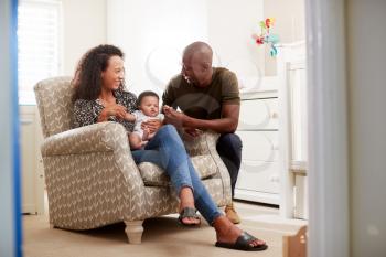 Loving Parents Sitting In Chair Cuddling Baby Son In Nursery At Home