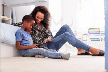 Mother With Son Sitting On Bed In Childs Bedroom Using Digital Tablet Together
