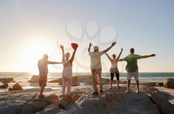 Rear View Of Senior Friends Standing On Rocks On Vacation With Arms Outstretched