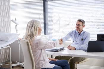 Mature Female Patient Shaking Hands With Doctor Sitting At Desk In Office