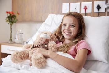 Portrait Of Girl Lying In Bed In Hospital Ward Hugging Teddy Bear