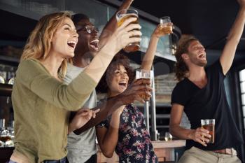 Group Of Male And Female Friends Celebrating Whilst Watching Game On Screen In Sports Bar