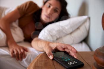 Mid adult woman waking up in bed, reaching out to smartphone on the bedside table in the foreground