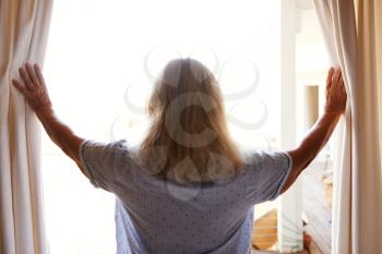 Smiling senior white woman opening the curtains on a sunny morning, back view, waist up