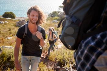 Young adult white man hiking with friends in countryside by the coast, smiling to camera