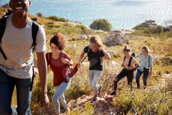 Young adult friends hiking single file uphill on a path by the coast, full length