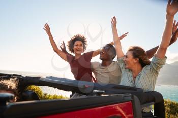 Excited millennial friends travelling in the back of an open car with their arms in the air