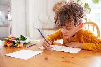 Young Boy At Home With Bunch Of Flowers Writing In Mothers Day Card