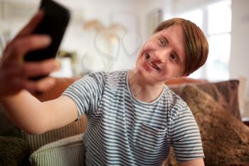 Young Downs Syndrome Man Sitting On Sofa Using Mobile Phone To Take Selfie At Home