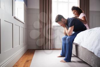 Woman Comforting Man Wearing Pajamas Suffering With Depression Sitting On Bed At Home