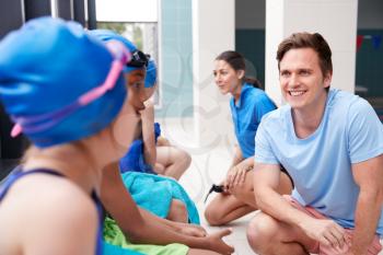 Male Coach Giving Children In Swimming Class Briefing As They Sit On Edge Of Indoor Pool