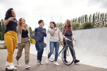 Female Friends With Skateboards And Bike Walking Through Urban Skate Park