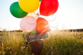 Rear View Of Two Female Friends Camping Running Through Field With Balloons Against Flaring Sun