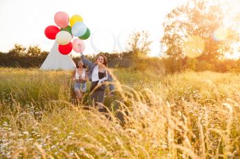 Portrait Of Two Female Friends Camping At Music Festival Running Through Field With Balloons