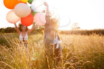 Two Female Friends Camping At Music Festival Running Through Field With Balloons Against Flaring Sun