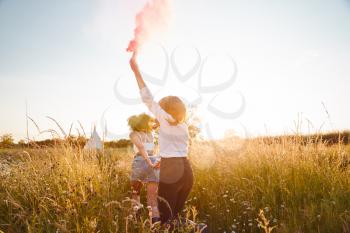 Rear View Of Two Female Friends Running Through Field With Smoke Flare