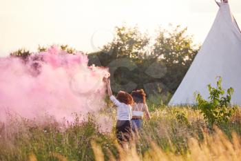 Rear View Of Two Female Friends Camping At Music Festival Running Through Field With Smoke Flare