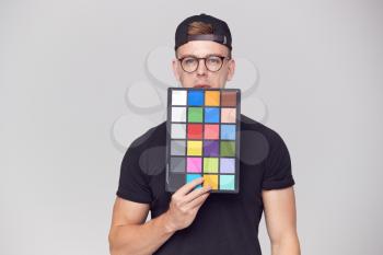Portrait Of Male Photographer In Studio Holding Colour Checker Card Against White Background