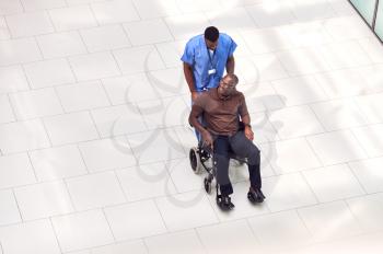 Overhead View Of Male Nurse Wheeling Patient In Wheelchair Through Lobby Of Modern Hospital Building