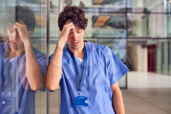 Tired Young Male Doctor Wearing Scrubs Leaning Against Wall In Modern Hospital Building