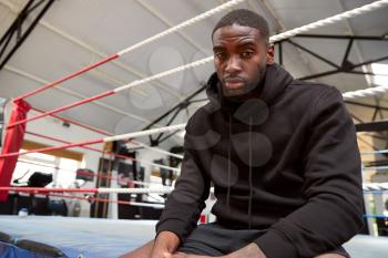 Portrait Of Male Boxer Wearing Hooded Sweatshirt In Gym Sitting On Boxing Ring