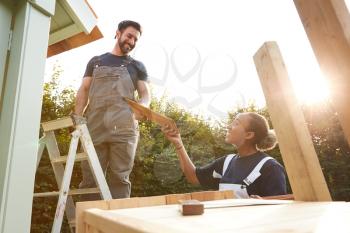 Male Carpenter With Female Apprentice Putting Roof On Outdoor Summerhouse In Garden