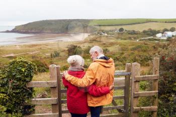 Rear View Of Active Senior Couple Looking Out Over Gate As They Walk Along Coastal Path In Fall