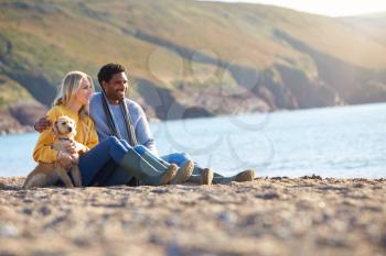 Loving Couple Sitting On Sand As They Walk With Dog Along Shoreline On Winter Beach Vacation