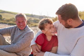 Family With Grandfather Relaxing On Beach With Adult Son And Grandson