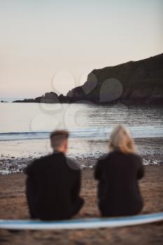 Defocused Shot Of Couple In Wetsuits On Surfing Staycation Sitting On Surfboard Looking Out To  Sea