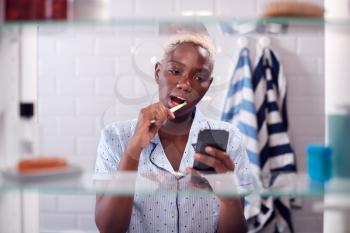 View Through Bathroom Cabinet Of Woman Brushing Teeth And Checking Phone Before Going To Work