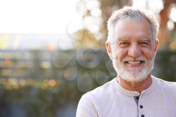 Portrait Of Smiling Retired Senior Hispanic Man In Garden At Home Against Flaring Sun