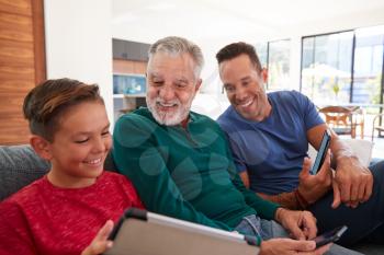 Multi-Generation Male Hispanic Family Sitting On Sofa At Home Using Mobile Phones And Digital Tablet