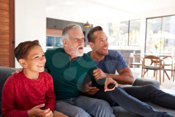 Excited Multi-Generation Male Hispanic Family Sitting On Sofa At Home Watching Sports On TV