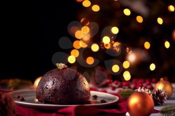 Traditional Christmas Pudding On Table Set For Festive Christmas Meal With Tree Lights In Background