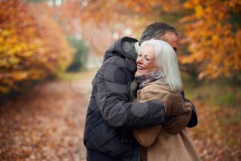 Loving Senior Couple Hugging As They Walk Along Autumn Woodland Path Through Trees