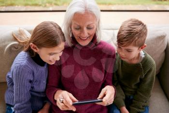 Grandchildren On Sofa At Home Showing Grandmother How To Use Mobile Phone