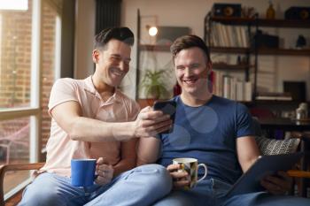 Loving Same Sex Male Couple Sitting On Sofa At Home Looking At Mobile Phone Together